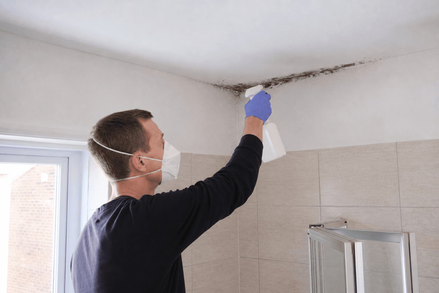 Homeowner in FFP3 mask spraying biocide on bathroom ceiling mould