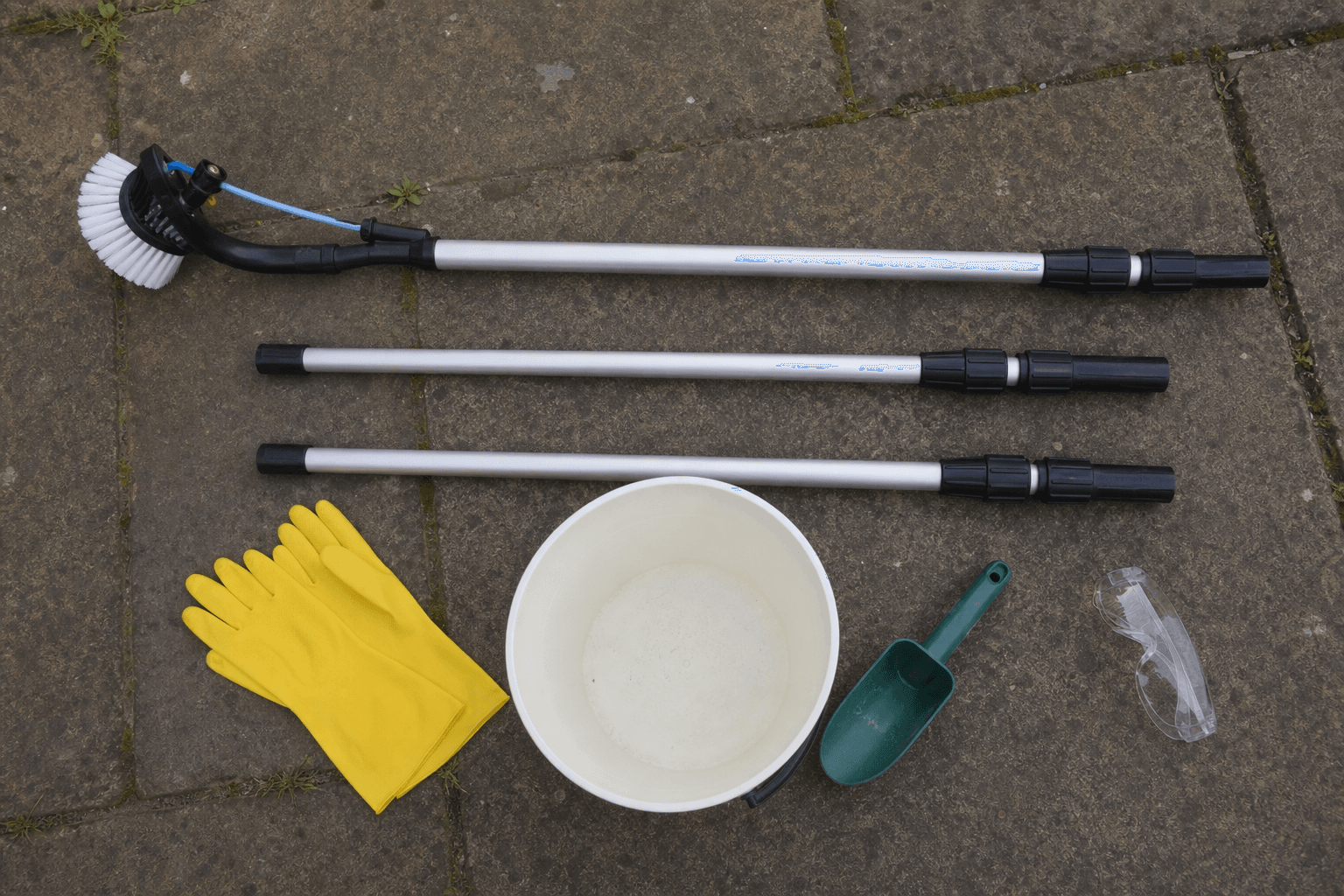 Flat-lay of UK gutter cleaning kit: telescopic pole, gloves, bucket, scoop and safety glasses on a patio