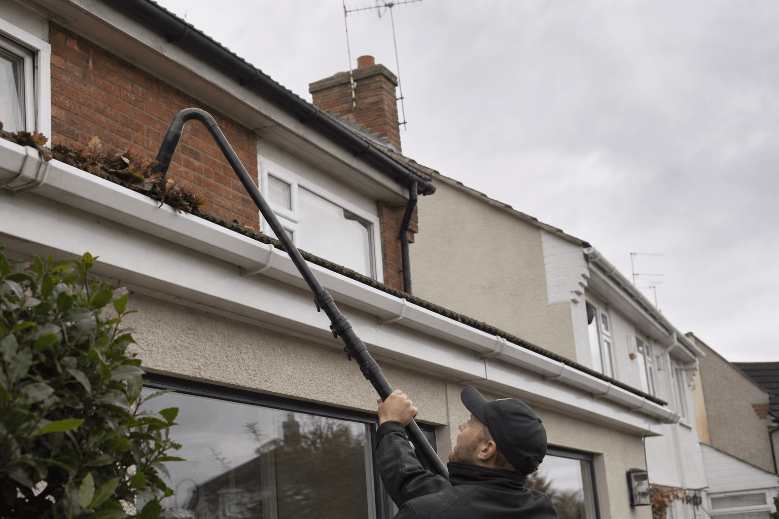 Person using a gutter vacuum on an extension pole at a UK terraced house, viewed from ground level